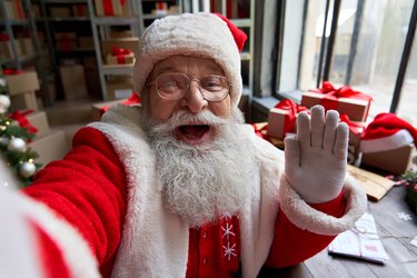 快乐的老胡子圣诞老人穿着服装手持电话挥手自拍、视频通话、录制视频圣诞快乐问候或拍摄 vlog 站在车间，面对相机视图。